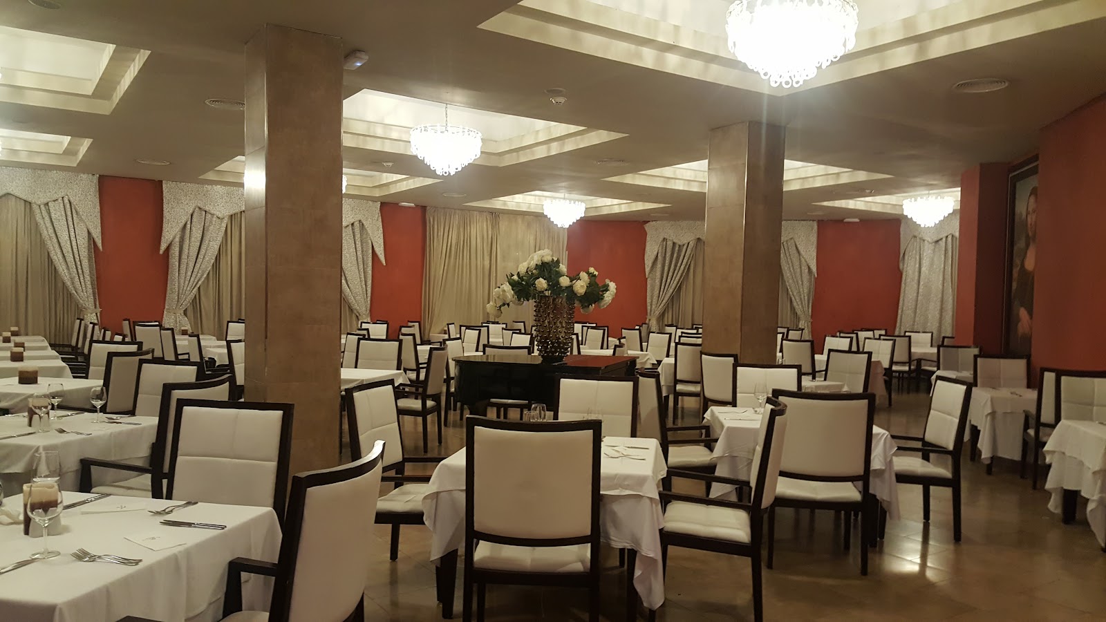 Formal dining room with white-clothed tables, cream and black chairs, red accent walls, crystal chandeliers, and a floral centerpiece.