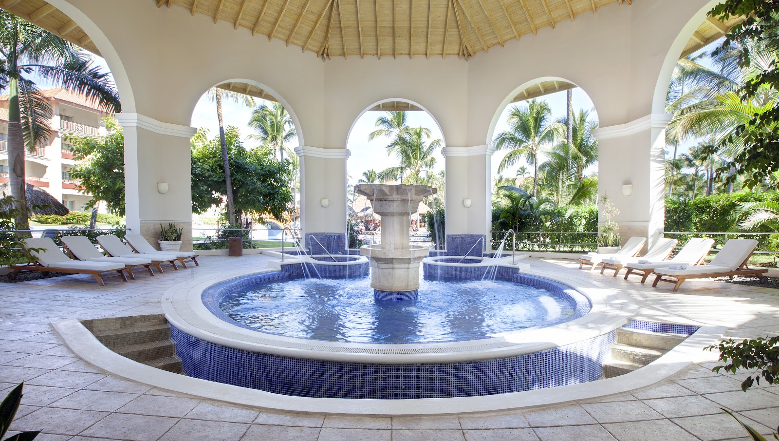Circular indoor pool with blue tile surround and central column, surrounded by white arches, lounge chairs, and palm trees visible through openings.