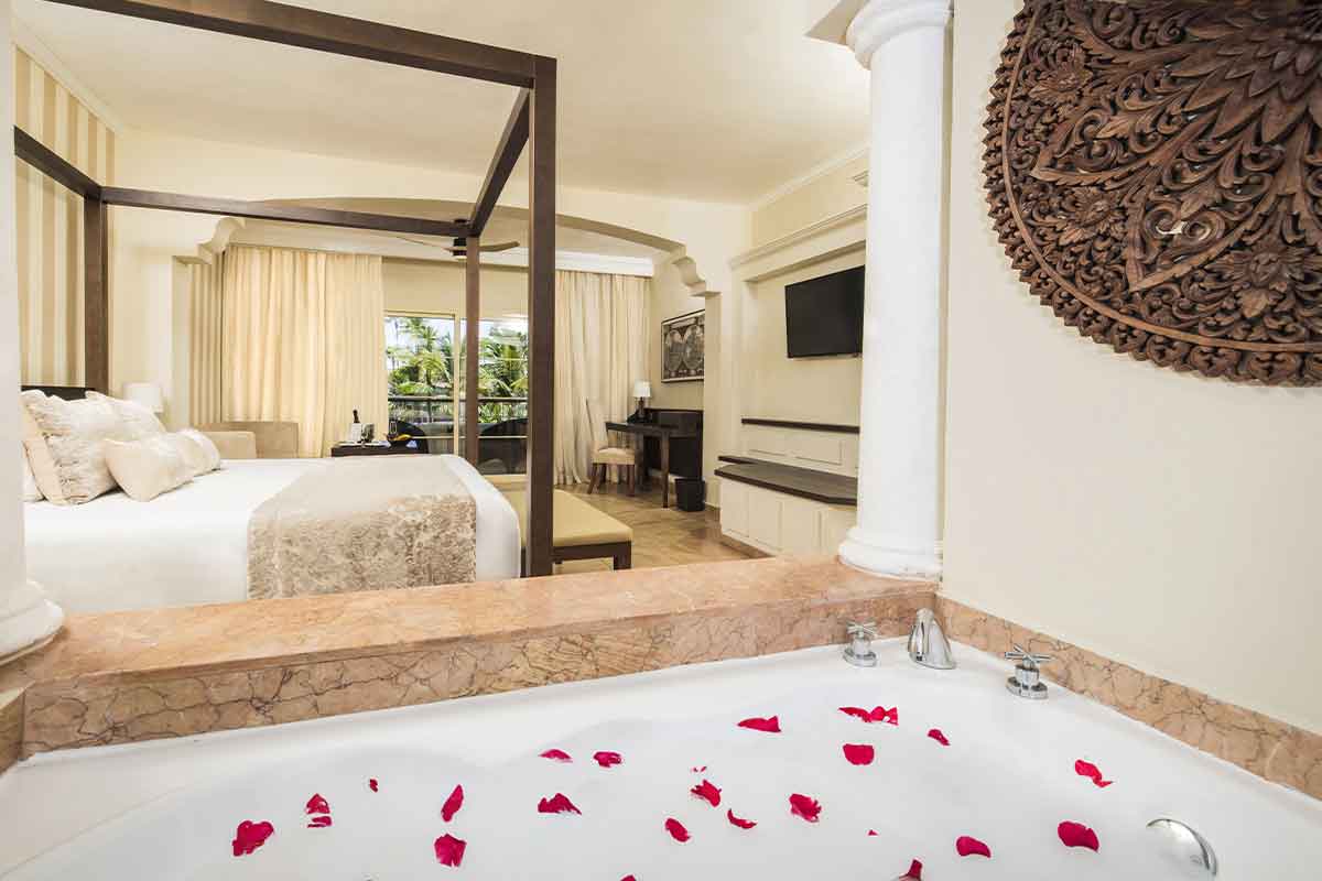 Bathtub scattered with red rose petals overlooking a spacious bedroom with four-poster bed and marble vanity at Majestic Colonial Punta Cana.