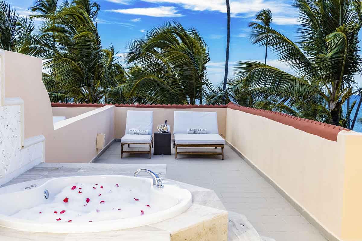 Private terrace with sunken tub filled with rose petals, two lounge chairs, and ocean view beyond swaying palms at Majestic Colonial Punta Cana.