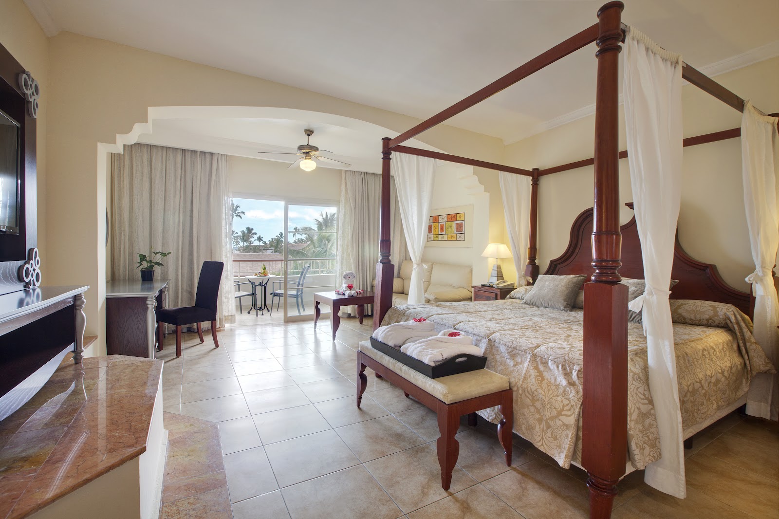 Spacious suite with four-poster bed, brown leather sofa, and sliding glass doors opening to a balcony with ocean view at Majestic Colonial Punta Cana.