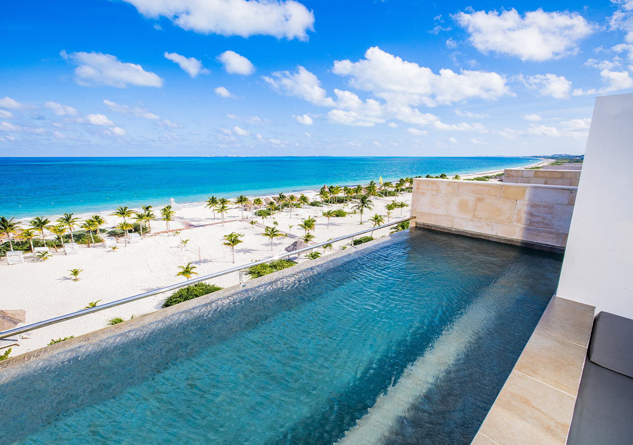 Resort pool overlooks white sand beach lined with palm trees and turquoise ocean under blue sky.
