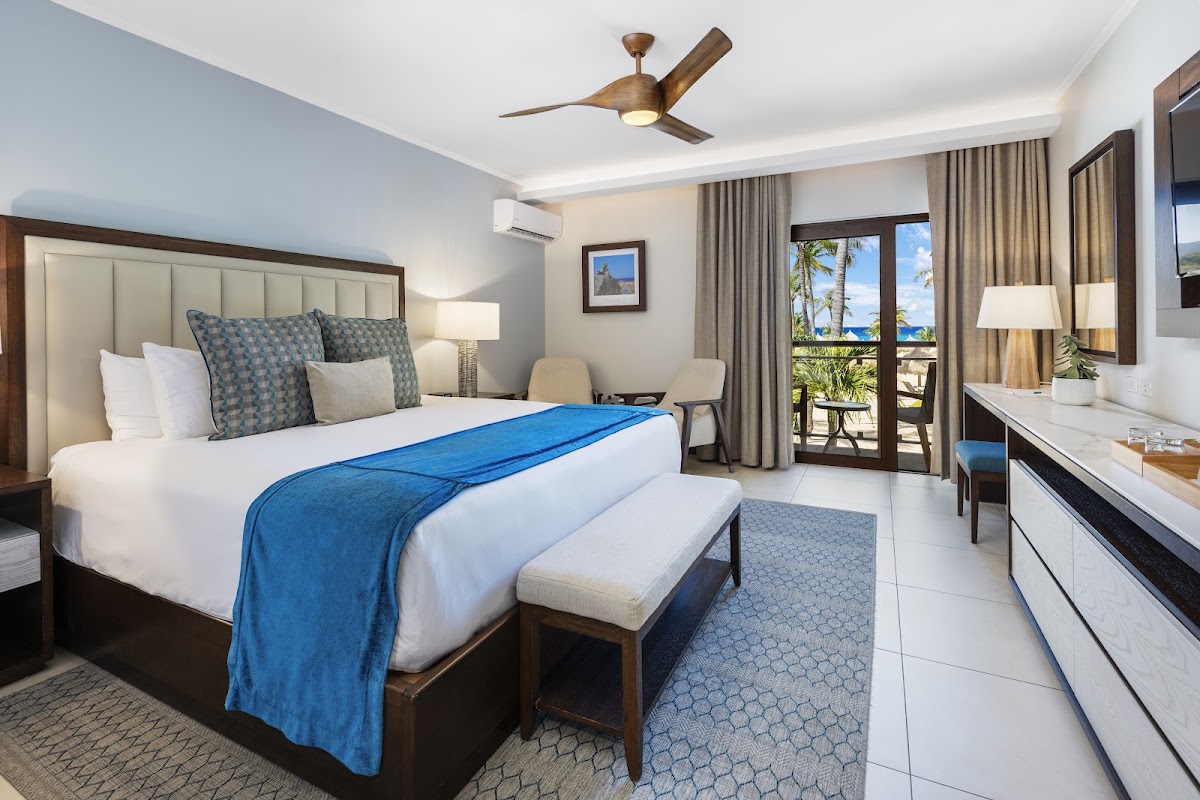 Resort bedroom with king bed draped in blue throw, ocean and palm tree view through balcony doors, ceiling fan, and vanity desk.
