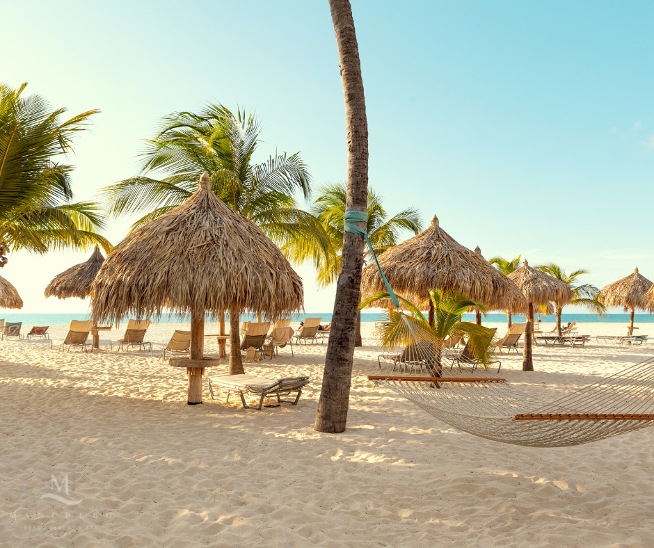 Beach area with thatched-roof palapas, lounge chairs, palm trees, and a hammock strung between palms on white sand.