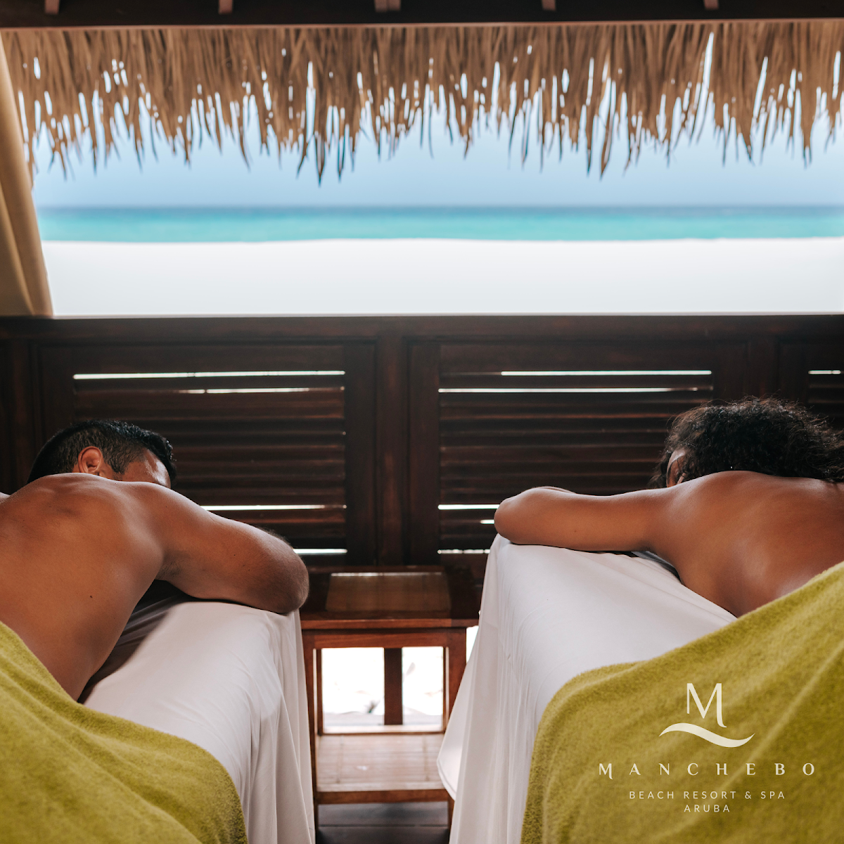 Two guests relax in lounge chairs under a thatched palapa at Manchebo Beach Resort & Spa, overlooking white sand and turquoise water.