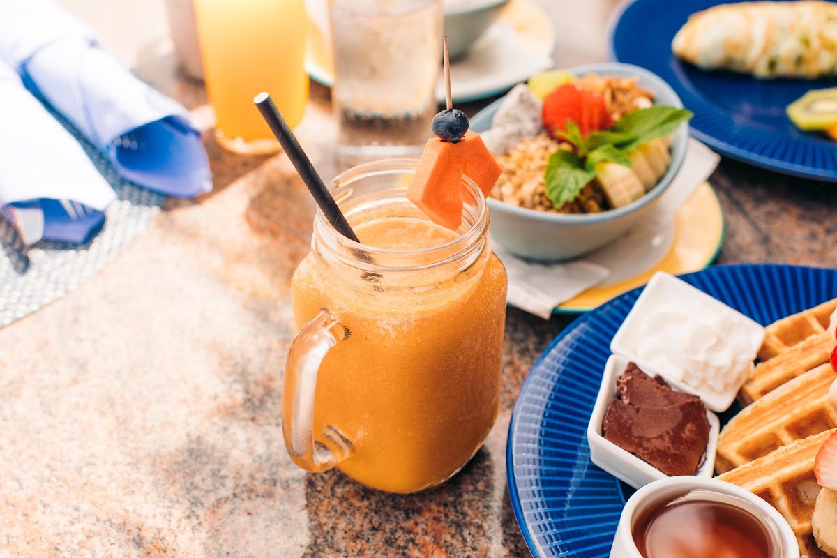 Orange cocktail in a mason jar with fruit garnish surrounded by blue plates of breakfast foods and juice on a stone table.