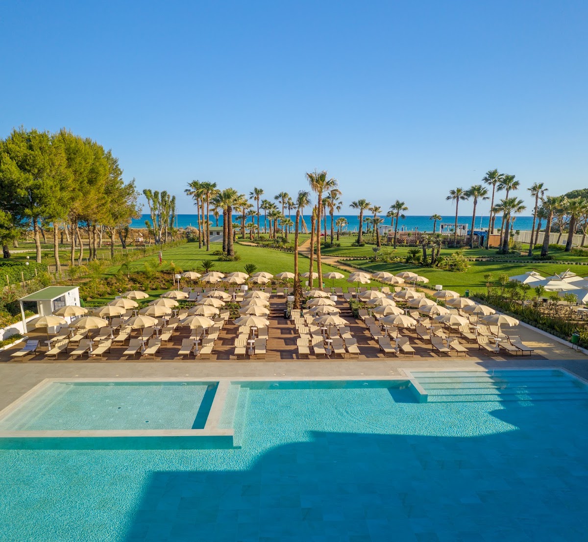 Resort pool deck with rows of tan umbrellas and loungers overlooking manicured grounds, palm trees, and Mediterranean Sea beyond.