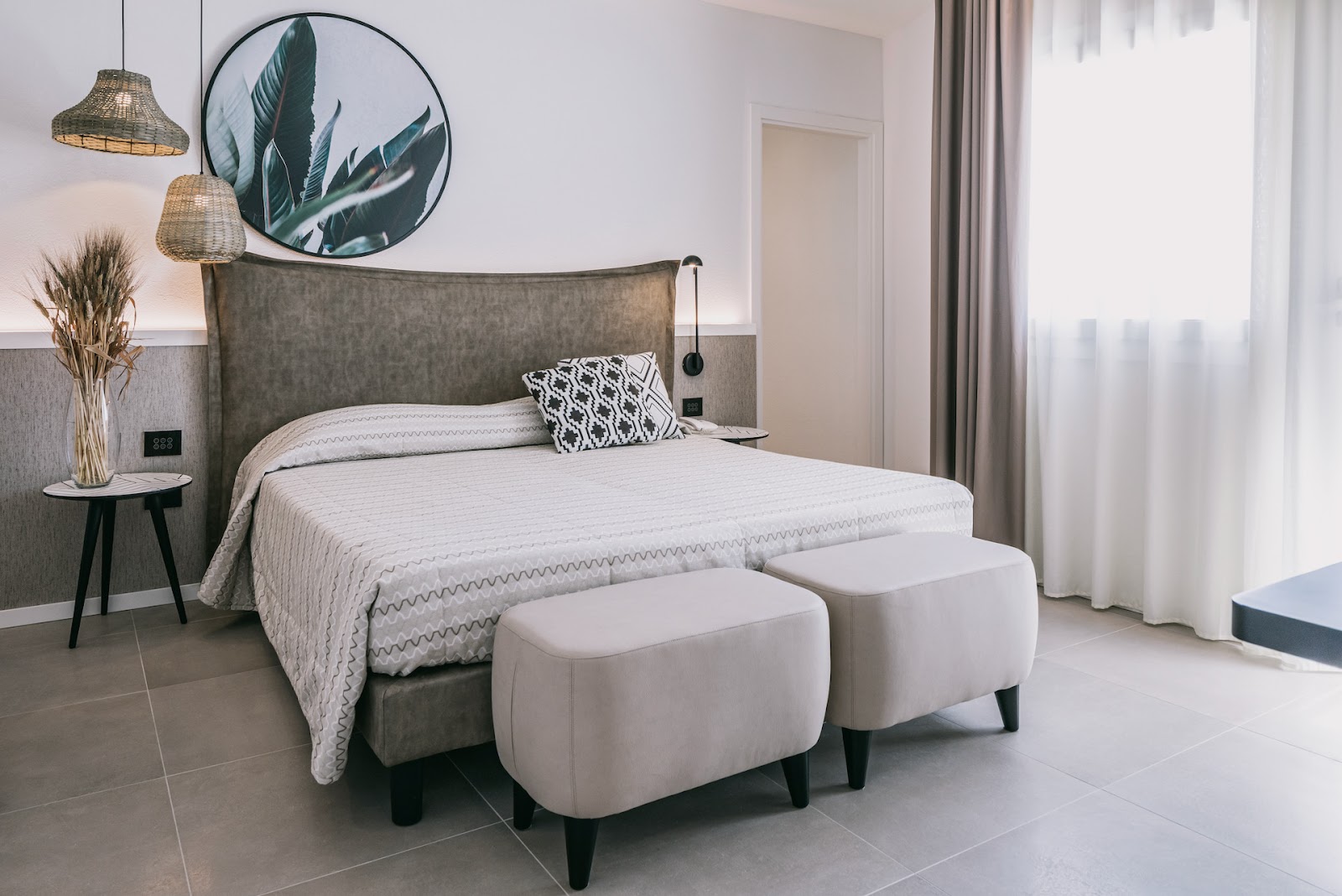 Modern resort bedroom with gray upholstered bed, white bedding, two cream ottomans, woven pendant lights, and circular botanical wall art.