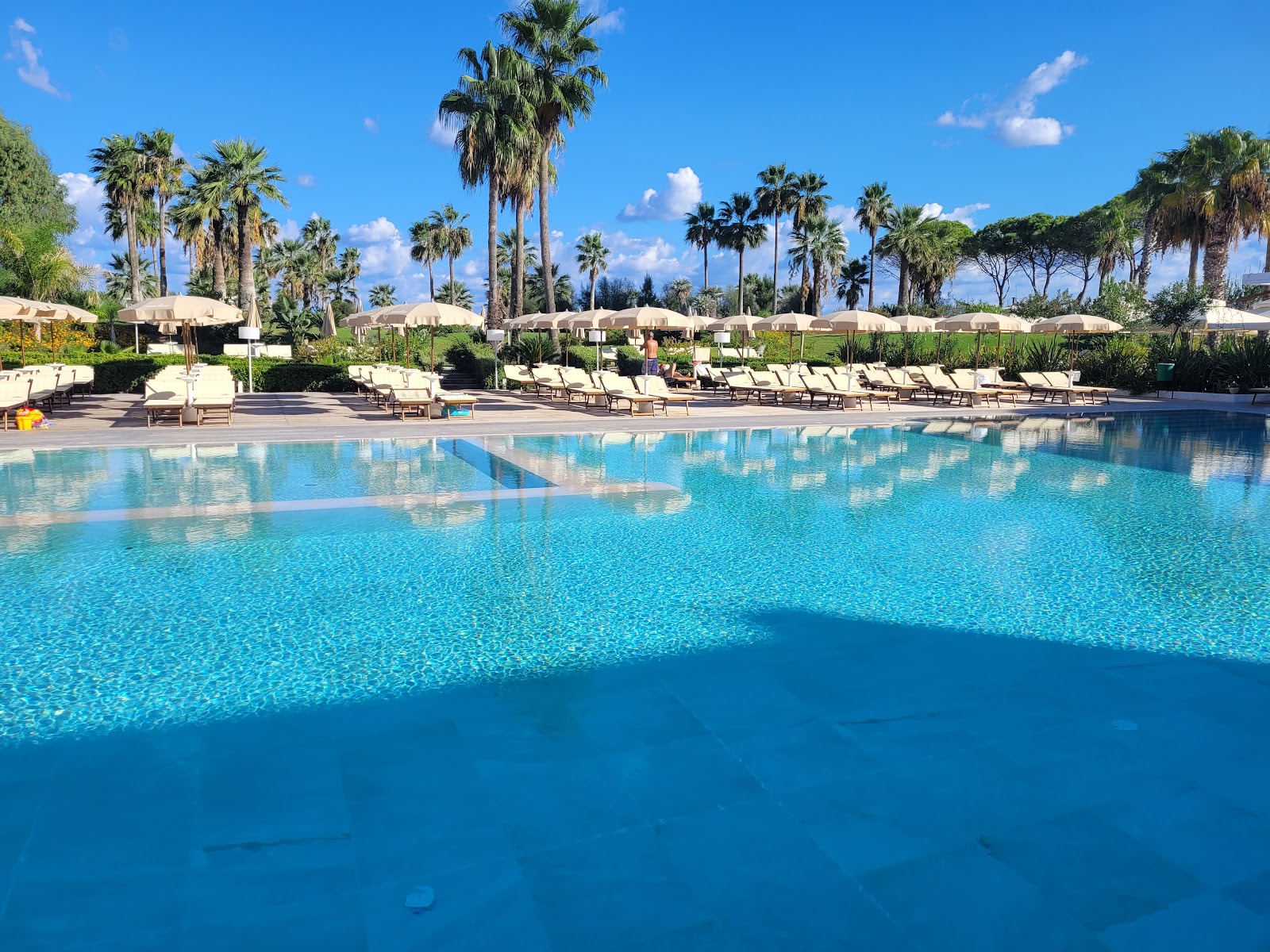 Large resort pool with clear blue water, bordered by beige lounge chairs and tan umbrellas, palm trees and green vegetation beyond under blue sky.