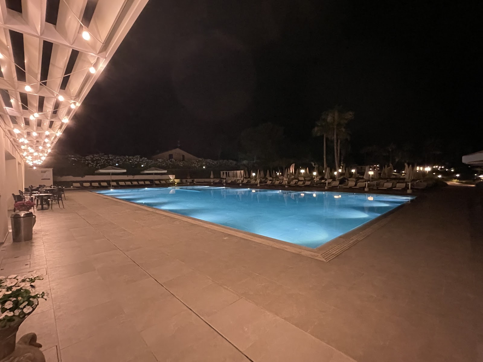 Illuminated pool at night with resort buildings, dining terrace, and palm trees along the deck at Mangia's Himera Resort.