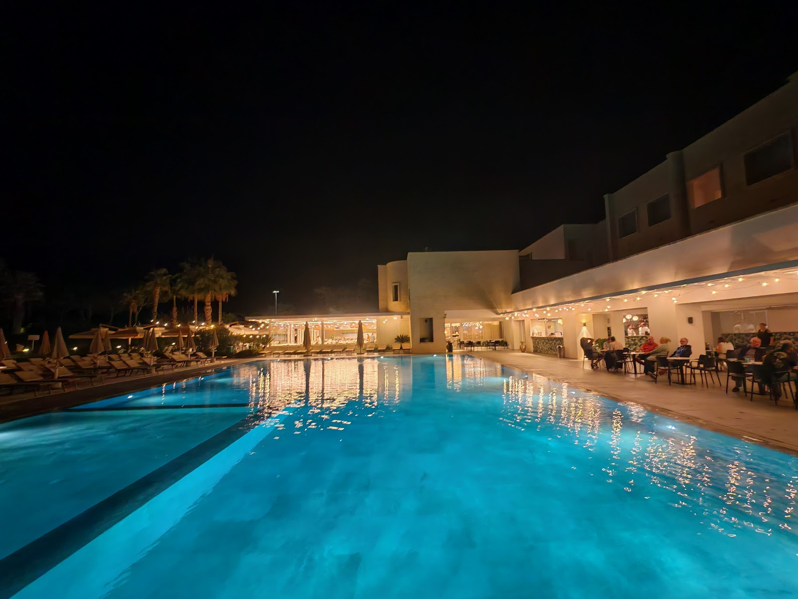 Beige resort buildings with palm-lined grounds overlook a sandy beach and turquoise Mediterranean Sea under clear sky.