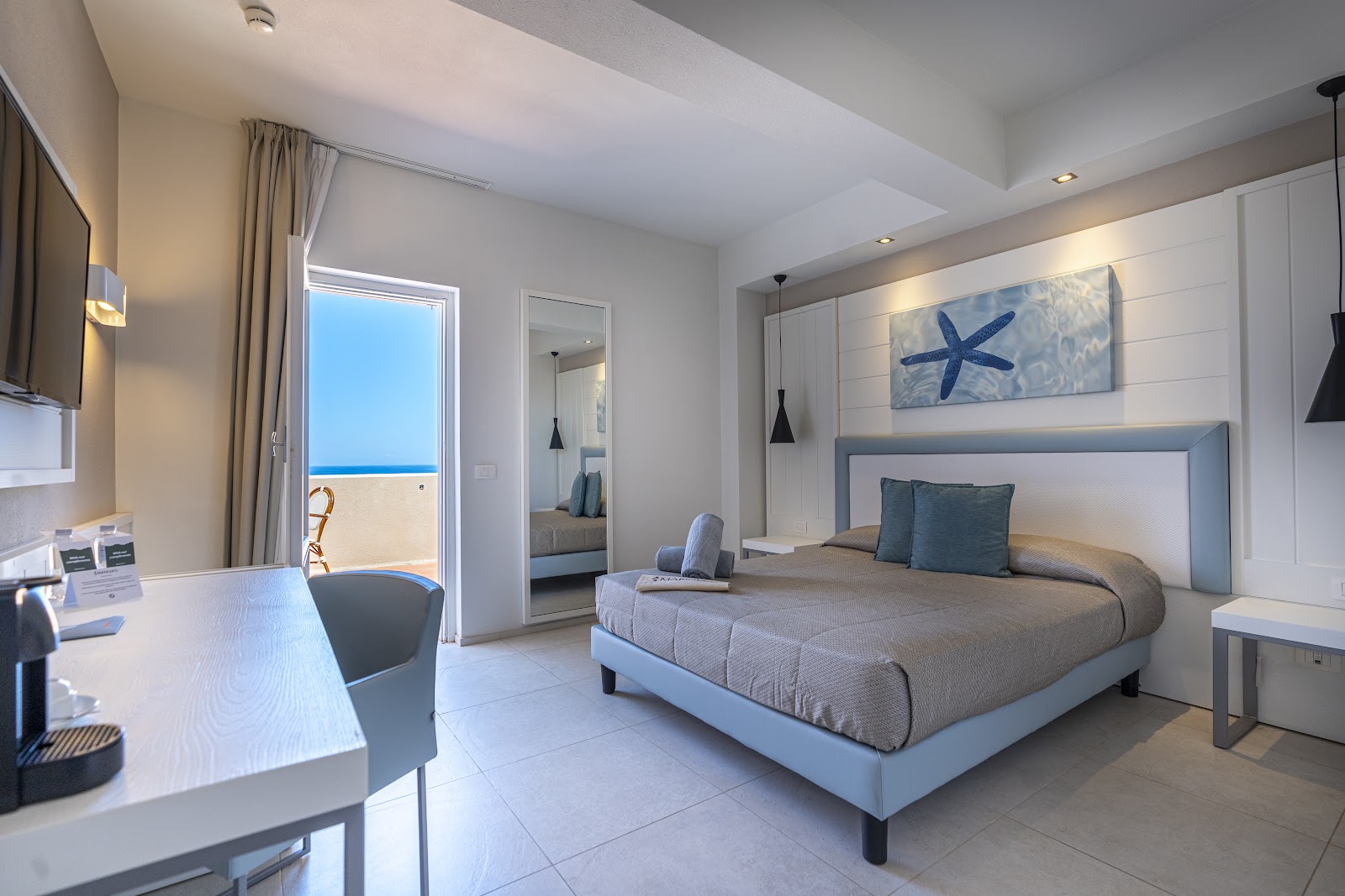 Modern resort bedroom with queen bed, starfish artwork, and ocean view through glass door to beachfront balcony.
