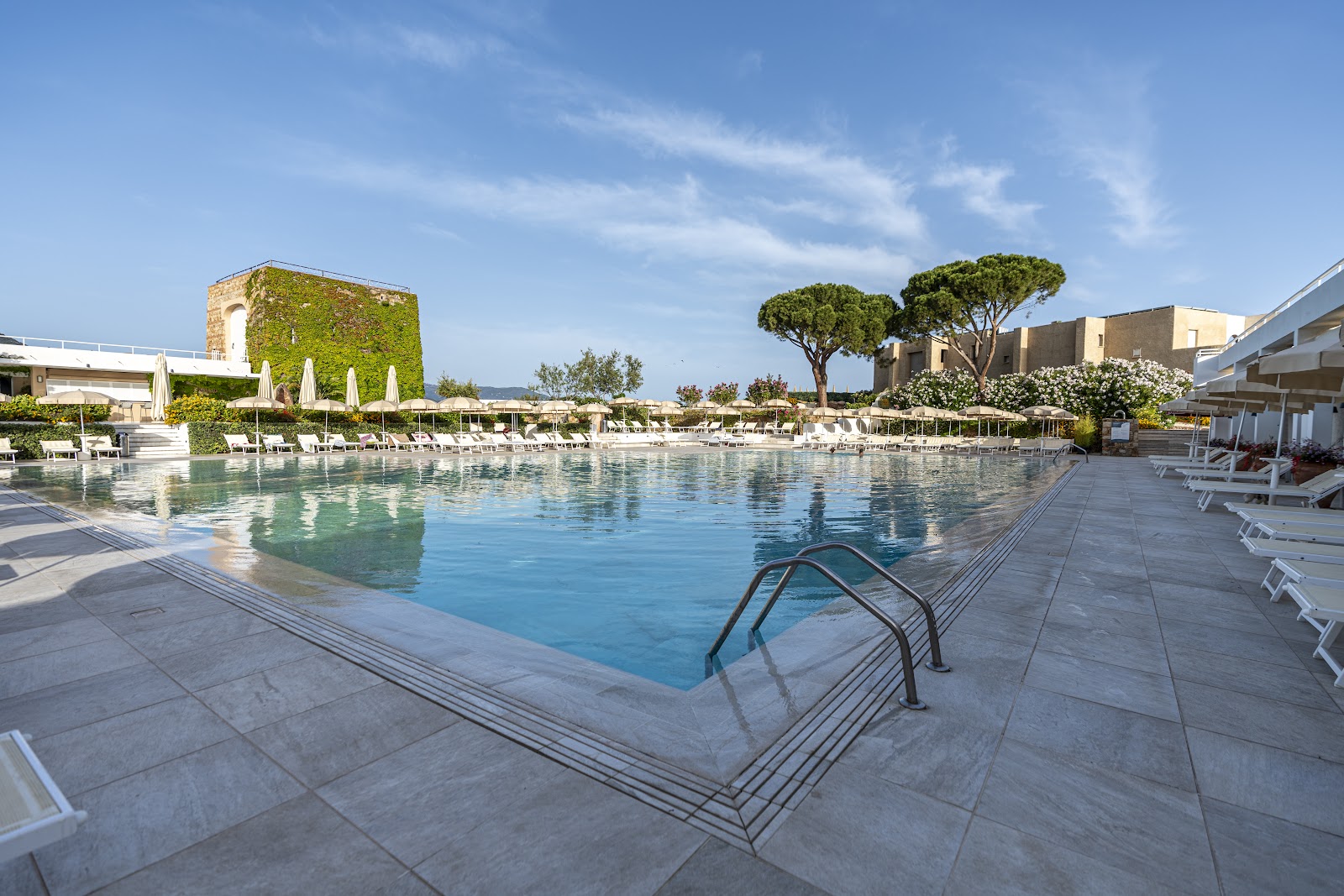 Resort pool with stone deck, white loungers, ivy-covered tower, and Mediterranean pine trees under blue sky.