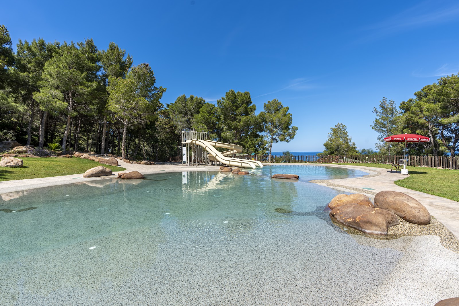 Resort pool with water slide, sandy beach entry, boulder landscaping, pine trees, and sea view under clear sky.