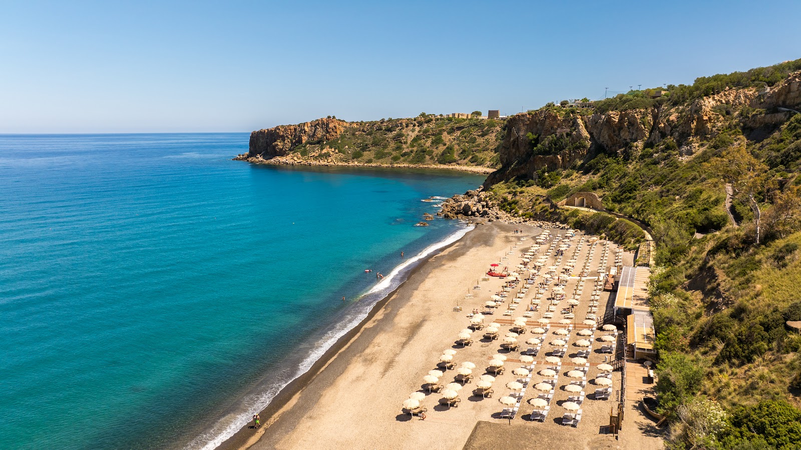 Sandy beach with rows of sun loungers backed by cliffs and turquoise Mediterranean waters at Mangia's Pollina Resort.