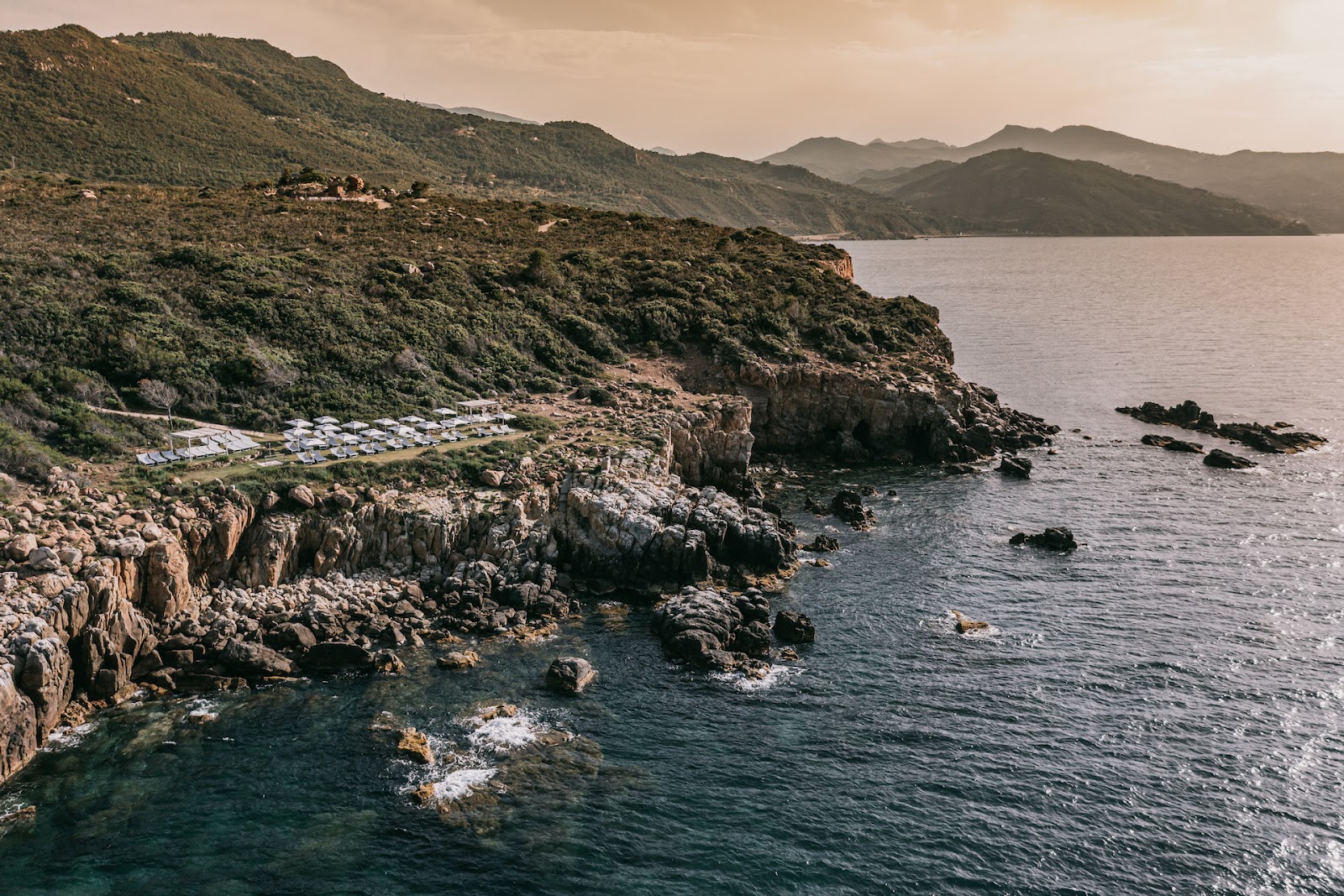 Wooden deck with yellow umbrellas and lounge chairs overlooking a Mediterranean bay with mountainous coastline.