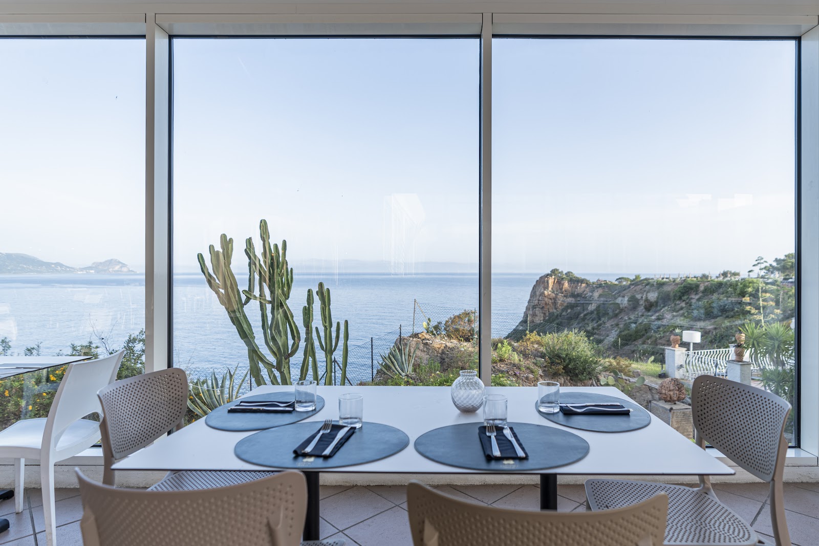 Clifftop terrace with white lounge seating, wooden railings, and potted plants overlooking blue Mediterranean Sea with distant islands.