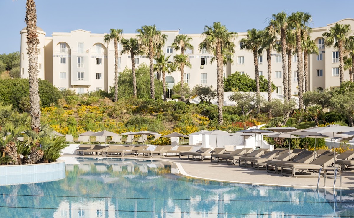 Cream-colored resort building with arched windows surrounded by palm trees, lounge chairs and umbrellas lining the pool deck with landscaped gardens beyond.