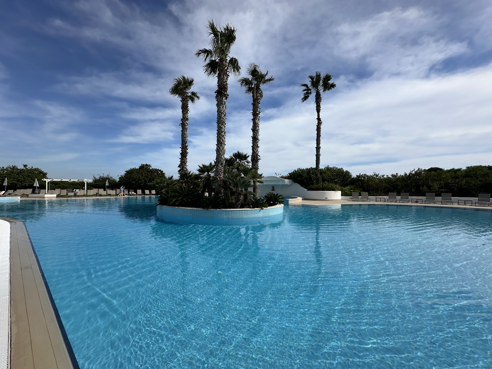 Water cascades into curved pool with loungers and umbrellas, cream-colored resort building and palm trees beyond.
