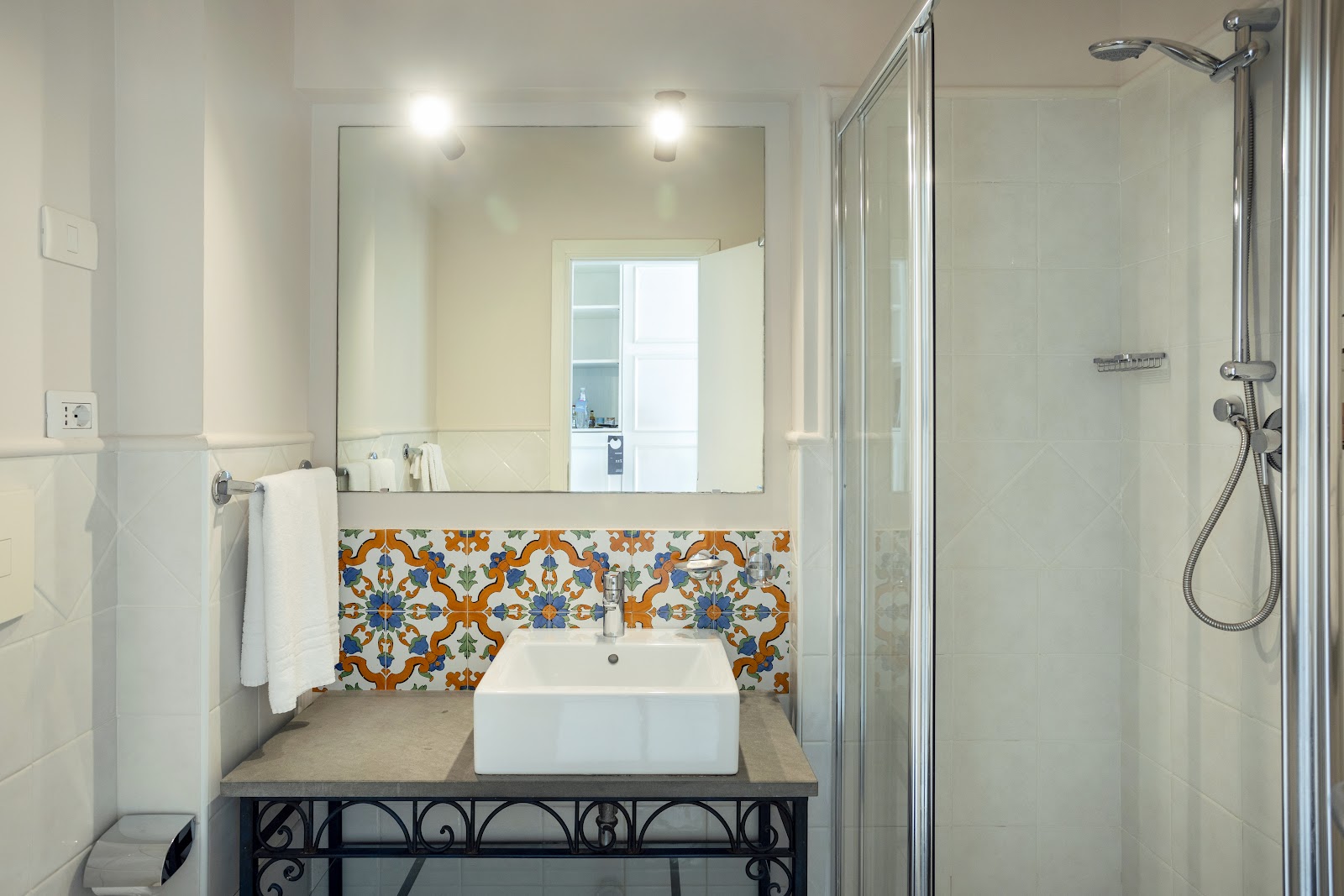 Modern bathroom with white rectangular sink on ornamental black vanity, decorative orange and blue tile backsplash, large mirror with sconces, and glass shower enclosure.