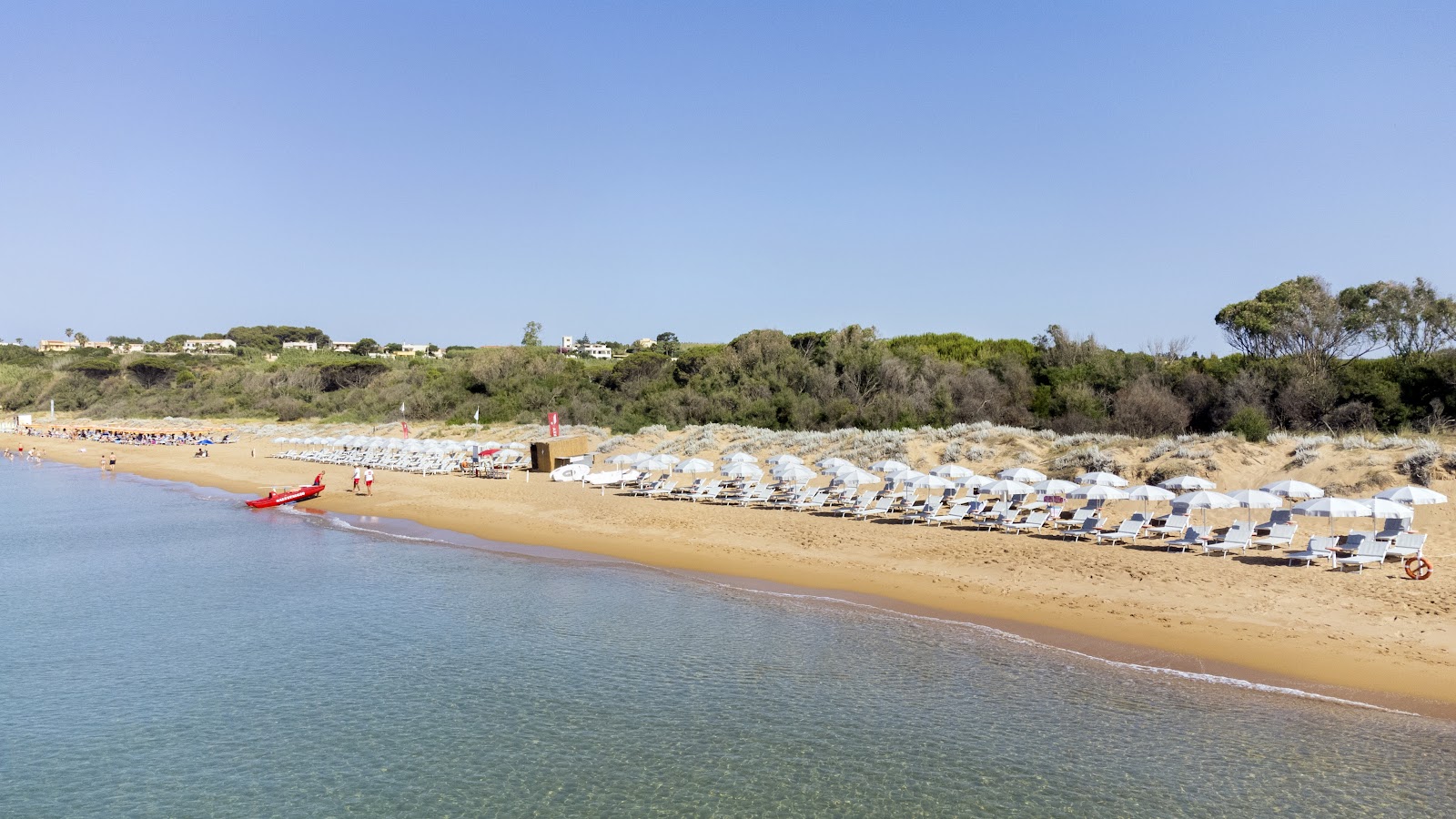 Sandy beach with organized rows of blue and white umbrellas and lounge chairs, calm waves, and green cliffs in the background.