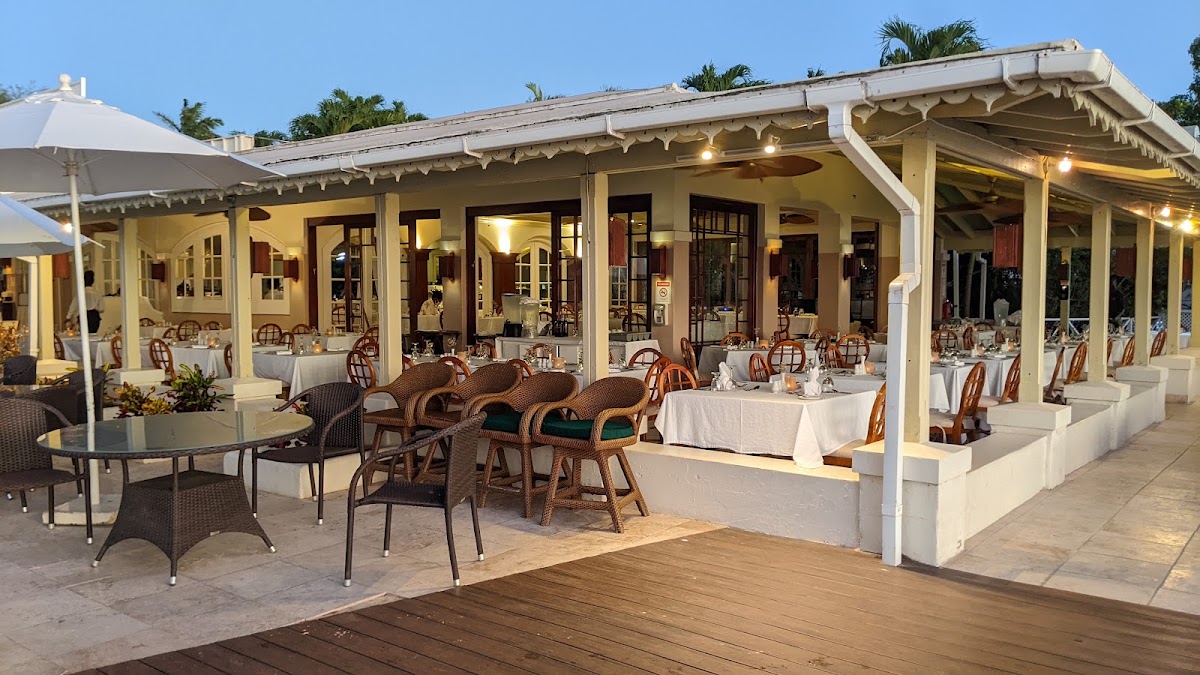 Cream-colored beachfront dining pavilion with white linens, woven chairs, and palm trees at dusk.