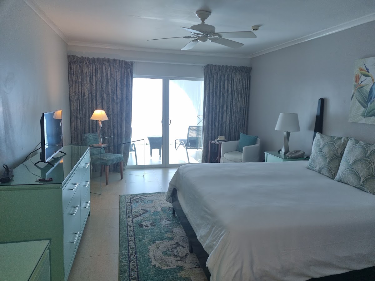 Resort hotel room with queen bed, pale blue walls, gray curtains, and glass doors opening onto a balcony overlooking the ocean.