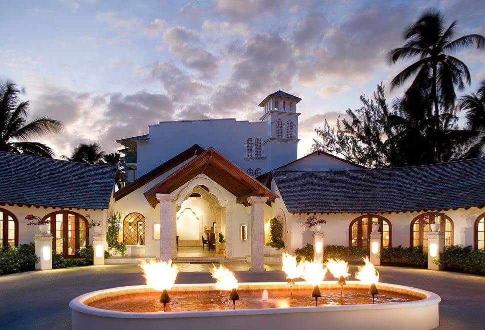 Illuminated white chapel with tower at dusk, flanked by lit arches, with lit fountain and palm trees in foreground at Mango Bay Hotel.