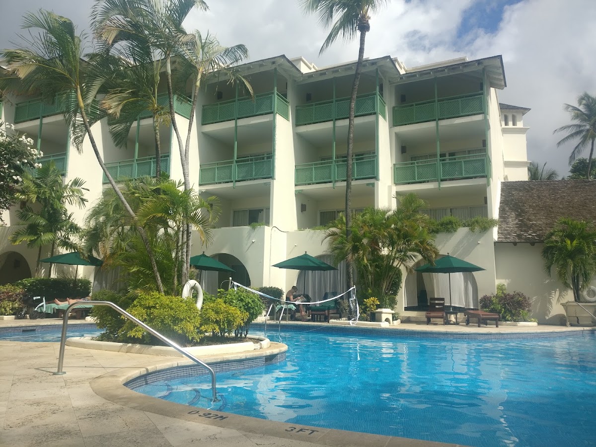 White multi-story hotel building with green balconies and palm trees beside a blue swimming pool with green umbrellas and lounge seating.