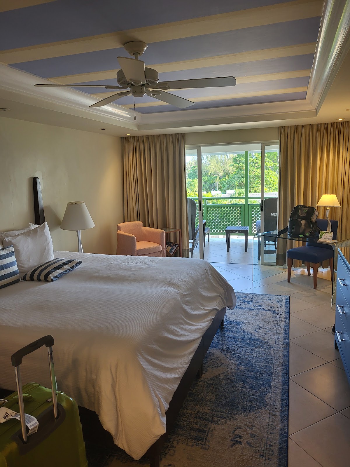 Spacious resort bedroom with queen bed, ceiling fan, and sliding glass doors opening to a balcony overlooking tropical greenery.