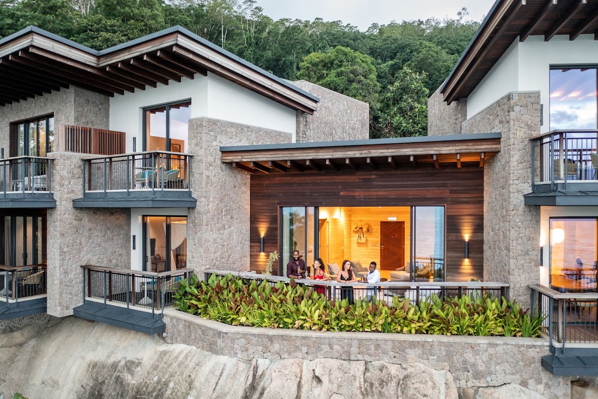 Modern stone and wood resort buildings with illuminated interiors, guests dining on a terrace overlooking tropical vegetation and rocky coastline at dusk.