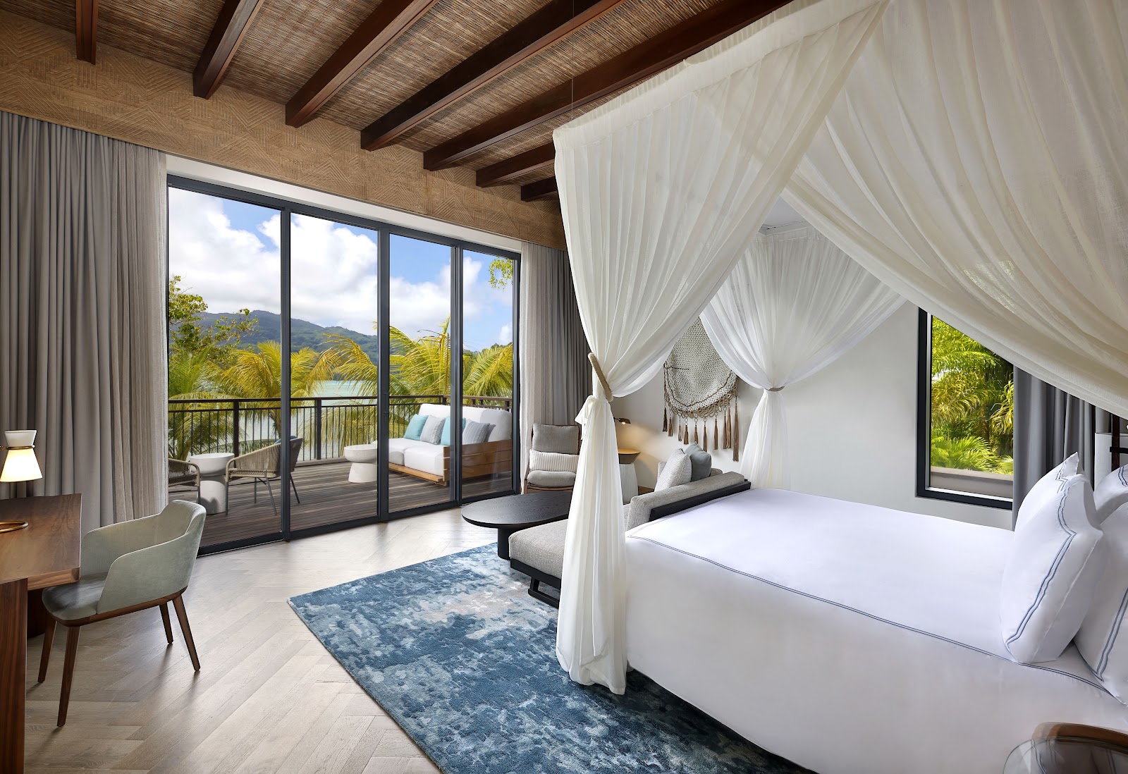 Spacious bedroom with canopied bed, wooden beams, and glass doors opening to a deck overlooking mountains and tropical vegetation at Mango House Seychelles.