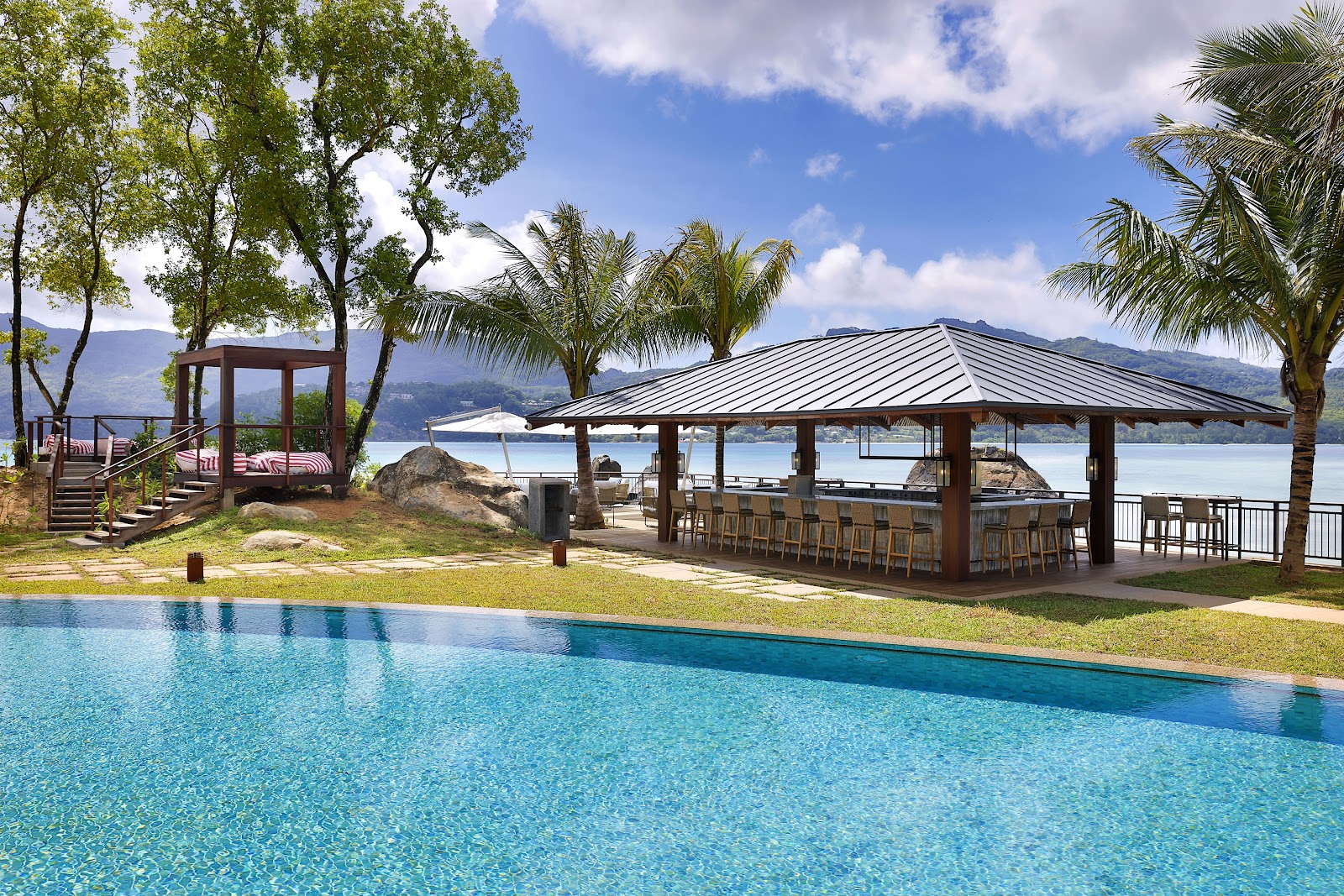 Clear pool in foreground with covered bar pavilion, wooden gazebo, and palm trees overlooking a calm bay and distant mountains.