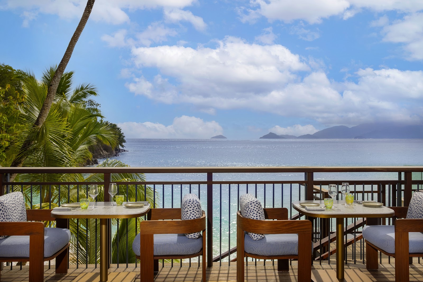 Dining tables on a beachfront deck at Mango House Seychelles with palm trees, calm ocean, and mountains visible across the water.