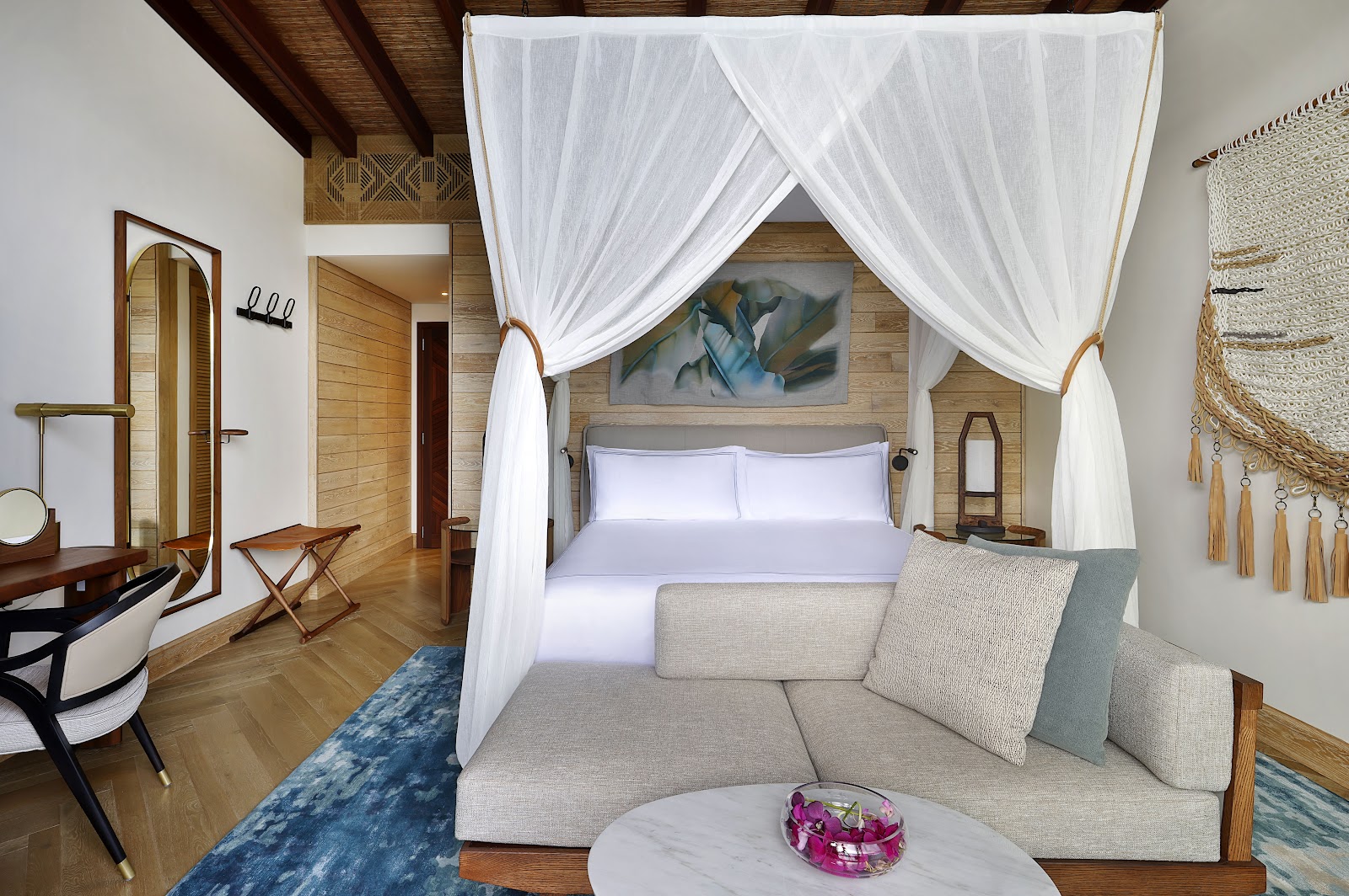 Resort bedroom with white canopy bed, gray upholstered sofa, wooden furnishings, and woven wall decor at Mango House Seychelles.