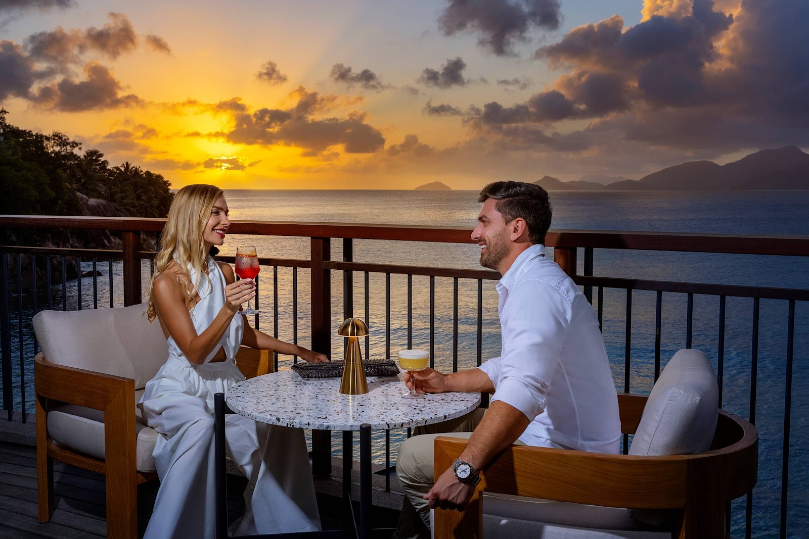 Couple toasts with cocktails at a seaside balcony table during sunset at Mango House Seychelles, with calm ocean and distant islands visible beyond the railing.