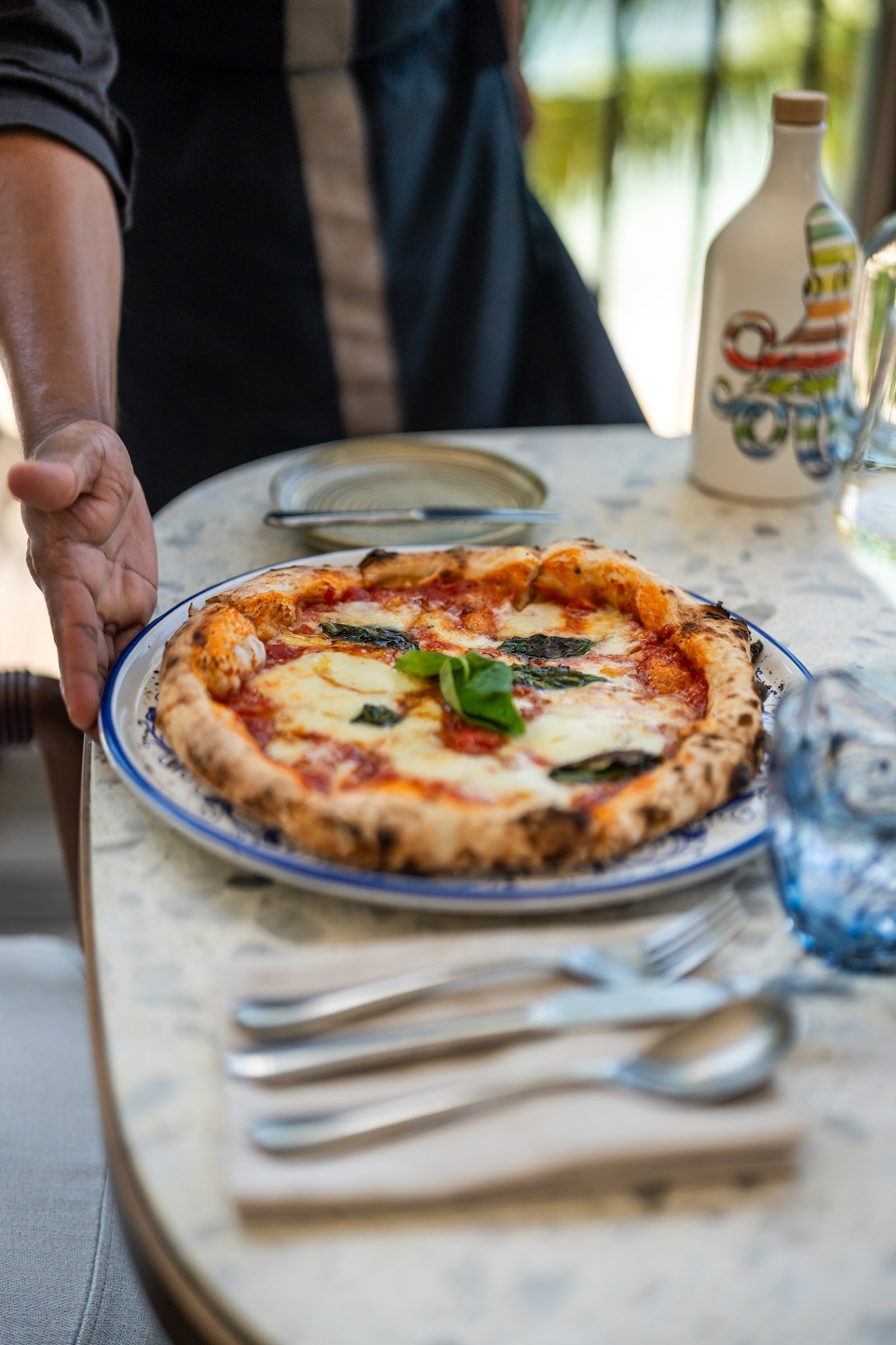 Person presenting a Margherita pizza with fresh basil at an outdoor dining table with water glasses and wine bottle visible.