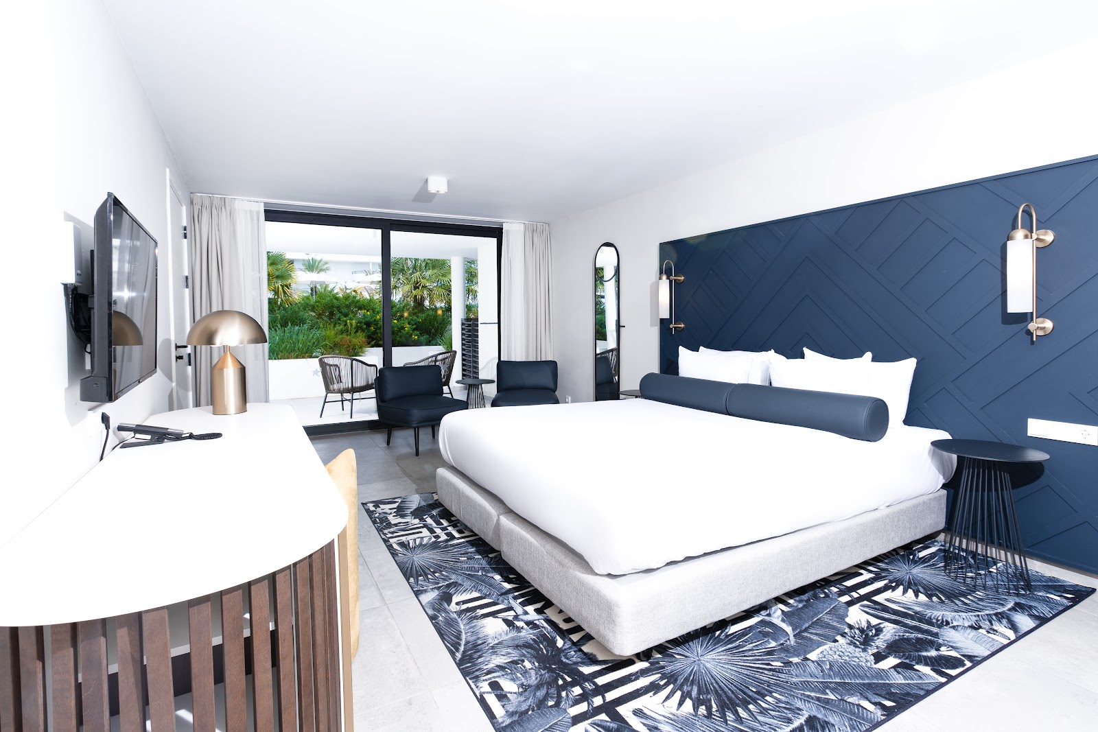 Modern resort bedroom with white bed, navy quilted headboard, oceanview balcony with tropical plants, and sitting area at Mangrove Beach Corendon Curaçao.