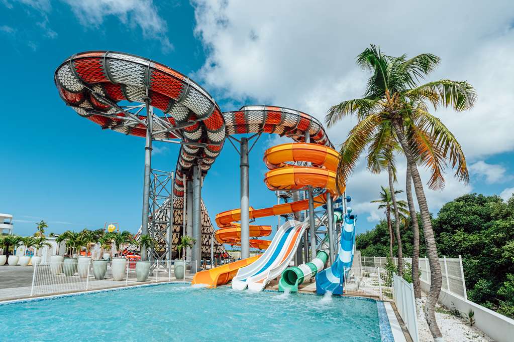 Water slides with red and orange tubes twist above a turquoise pool surrounded by palm trees at Mangrove Beach Corendon Curaçao.