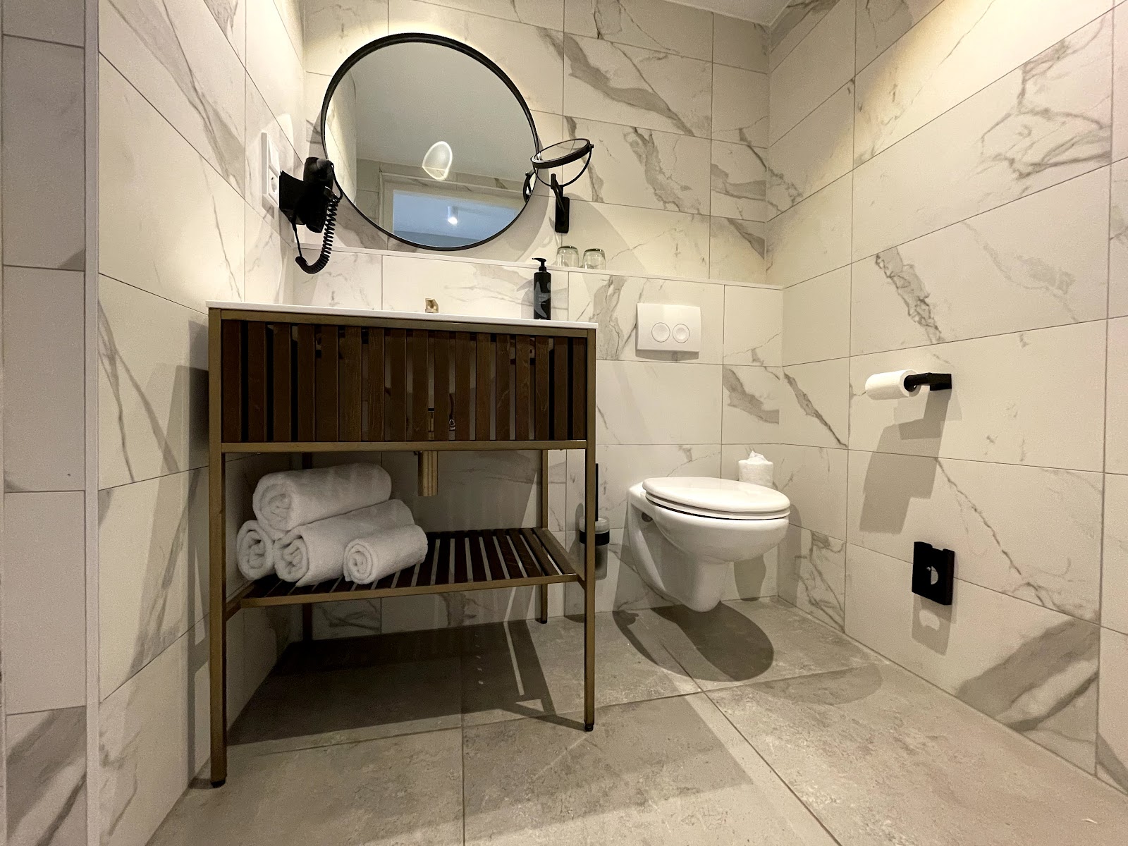 Modern bathroom with marble-look tiles, round mirror with black frame, wooden vanity holding rolled towels, and white toilet.