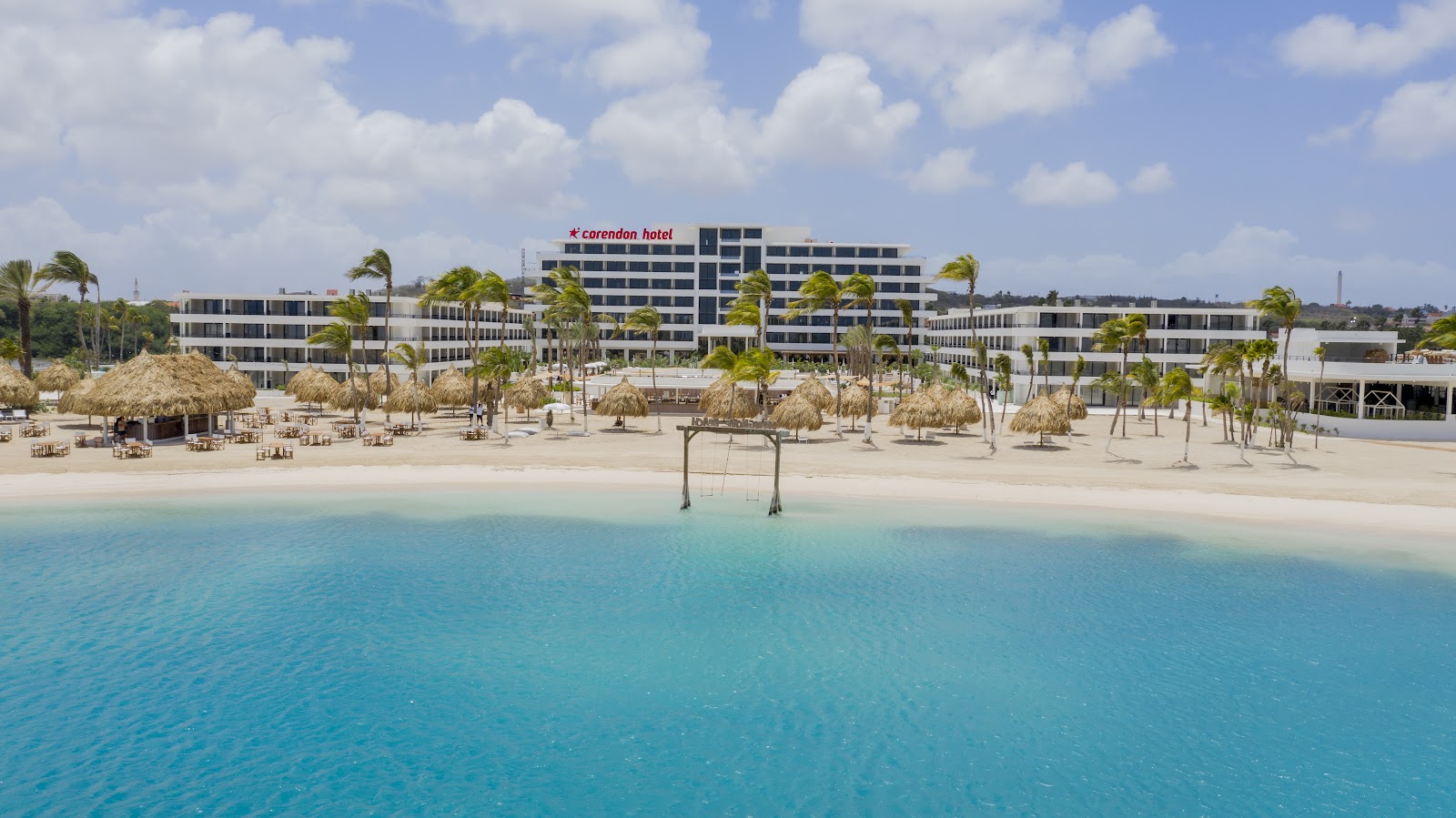 Corendon Hotel overlooks a sandy beach with thatched palapas, palm trees, and turquoise water under a cloudy sky.