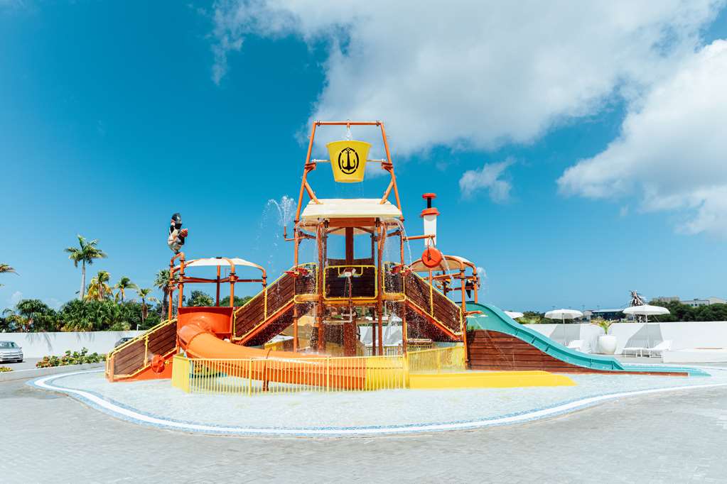 Pirate ship-themed water play structure with orange and yellow slides at Mangrove Beach Corendon Curaçao, surrounded by shallow pool water under blue sky.
