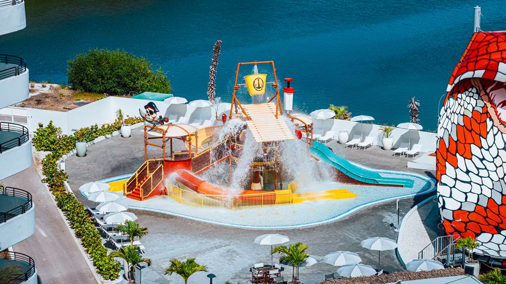 Overhead view of a water park with colorful slides, splash features, and a large red dragon sculpture at Mangrove Beach Corendon Curaçao, surrounded by white loungers and turquoise ocean.