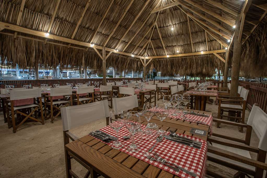 Open-air dining pavilion with thatched palm roof, wooden tables set with red and white checkered cloths, and water visible beyond at Mangrove Beach Corendon Curaçao.