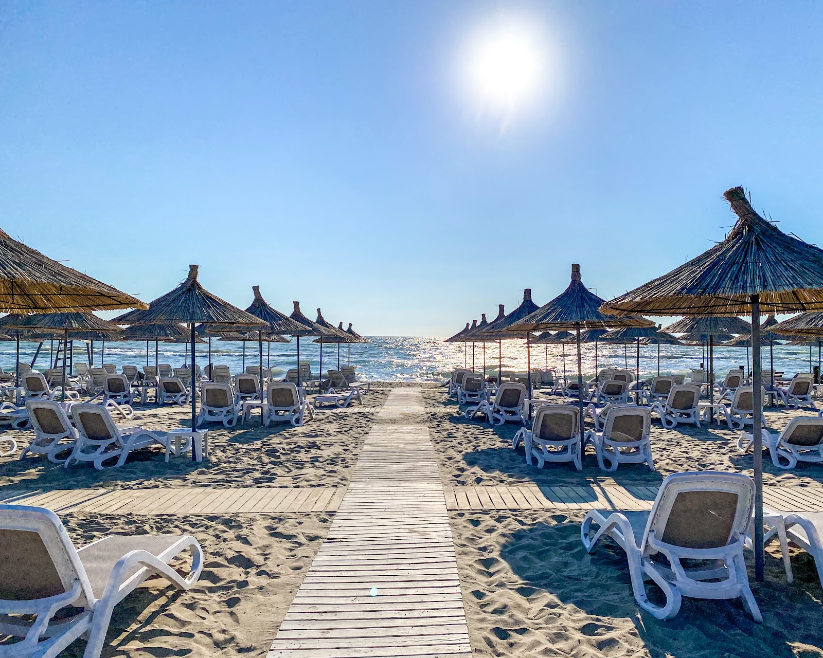 Wooden boardwalk leading through rows of white loungers under thatched umbrellas on a sandy beach with sparkling blue water under bright sun.