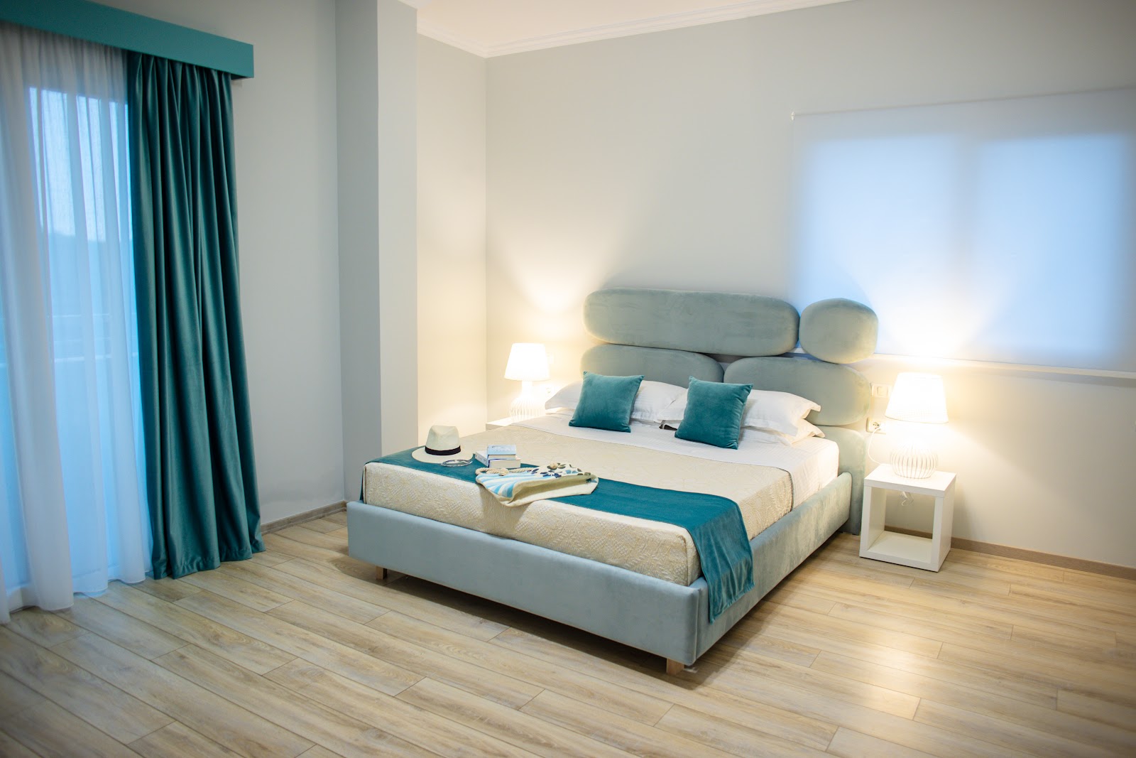 Bright bedroom with white walls, cream bed with teal pillows and headboard, teal curtains, wood flooring, and bedside lamps at MarBella Beach Hotel.