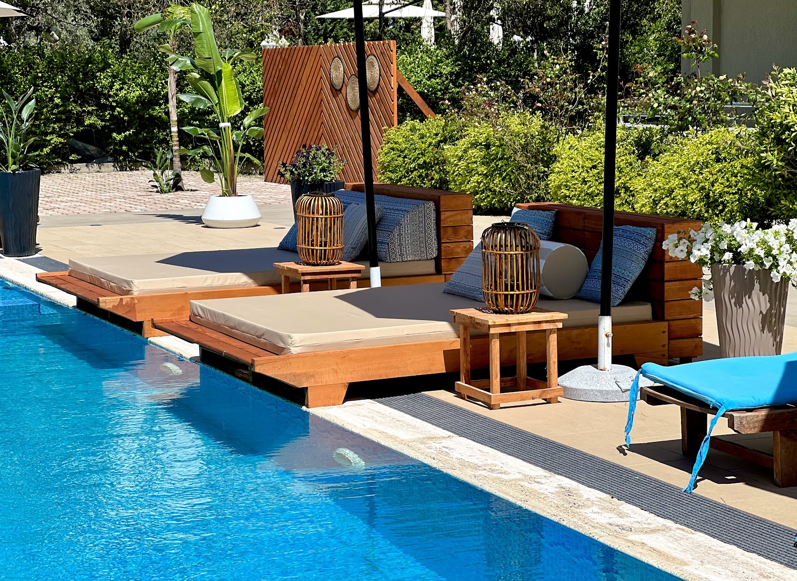 Pool deck at MarBella Beach Hotel with wooden loungers, woven lanterns, and lush greenery bordering the blue water.