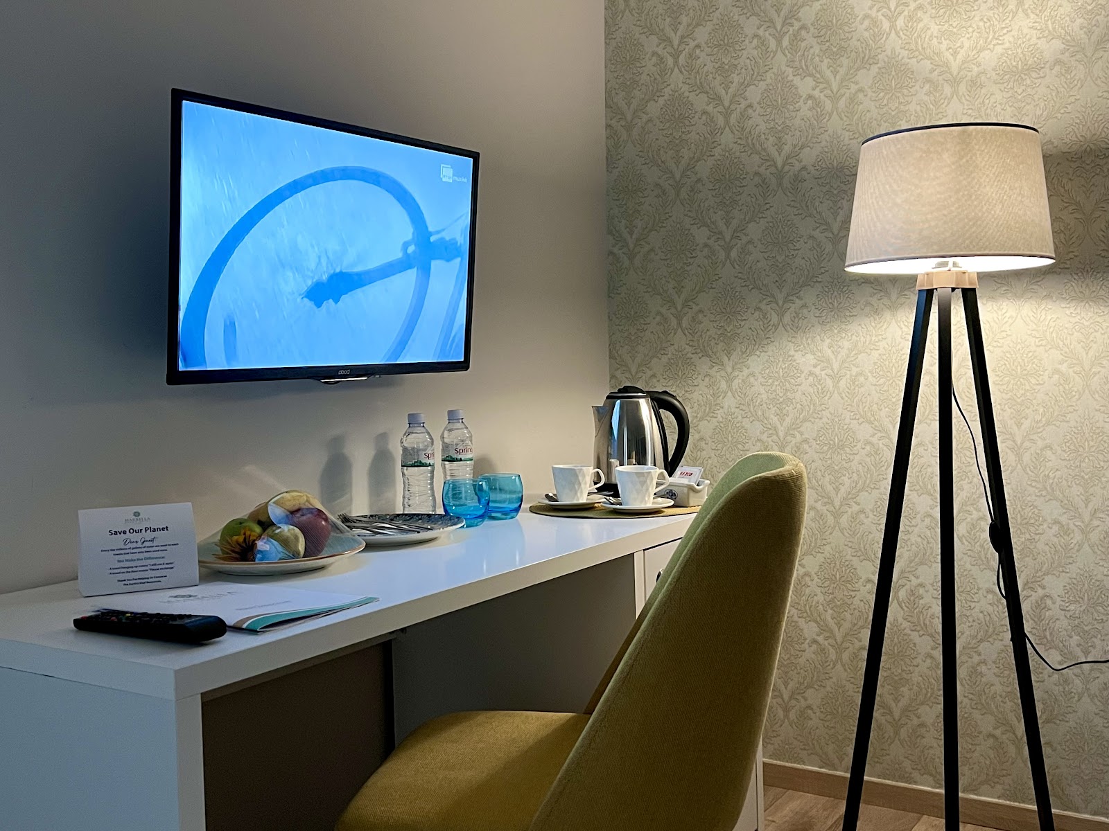 Guest room desk with water bottles, fruit plate, kettle, and wall-mounted TV showing a helicopter, tripod floor lamp nearby.