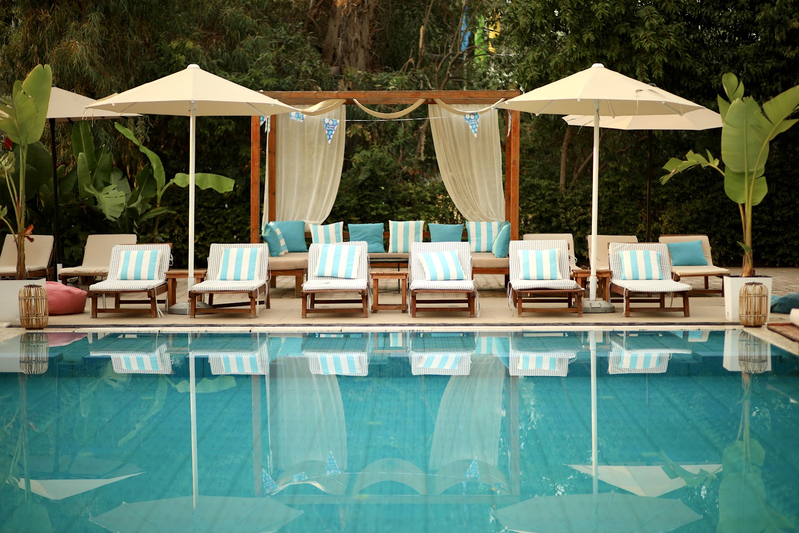 Turquoise pool bordered by striped lounge chairs under cream umbrellas and a draped pergola, with tropical plants and dense greenery behind.