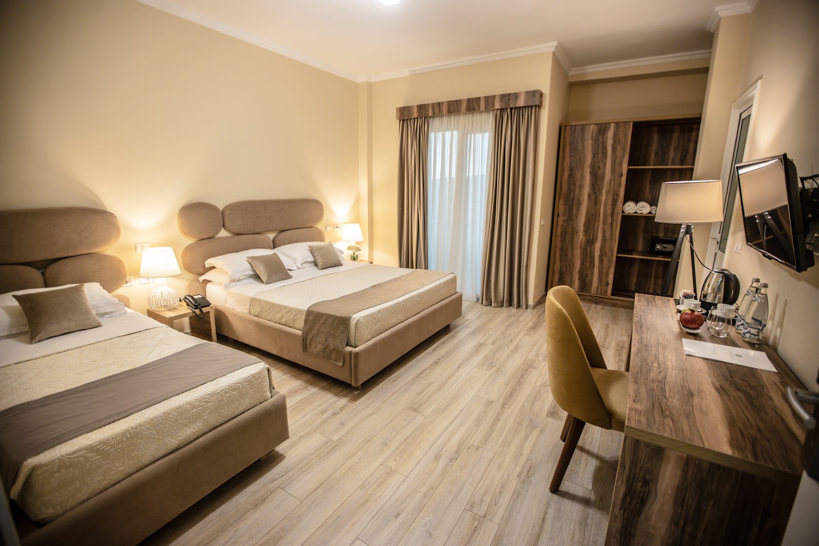 Spacious resort bedroom with two beds, wooden desk with chair, TV, wood-grain flooring, and balcony access at MarBella Beach Hotel.